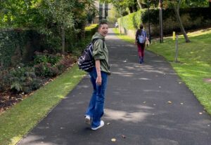 A photo of Lois walking along a tree-lined path, looking over their shoulder at the camera and smiling widely