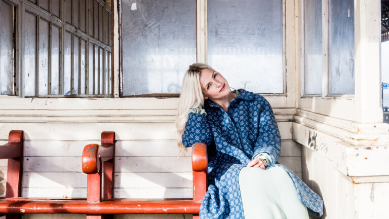 photo of Jenefer Odell sitting on a bench, wearing a blue coat and smiling