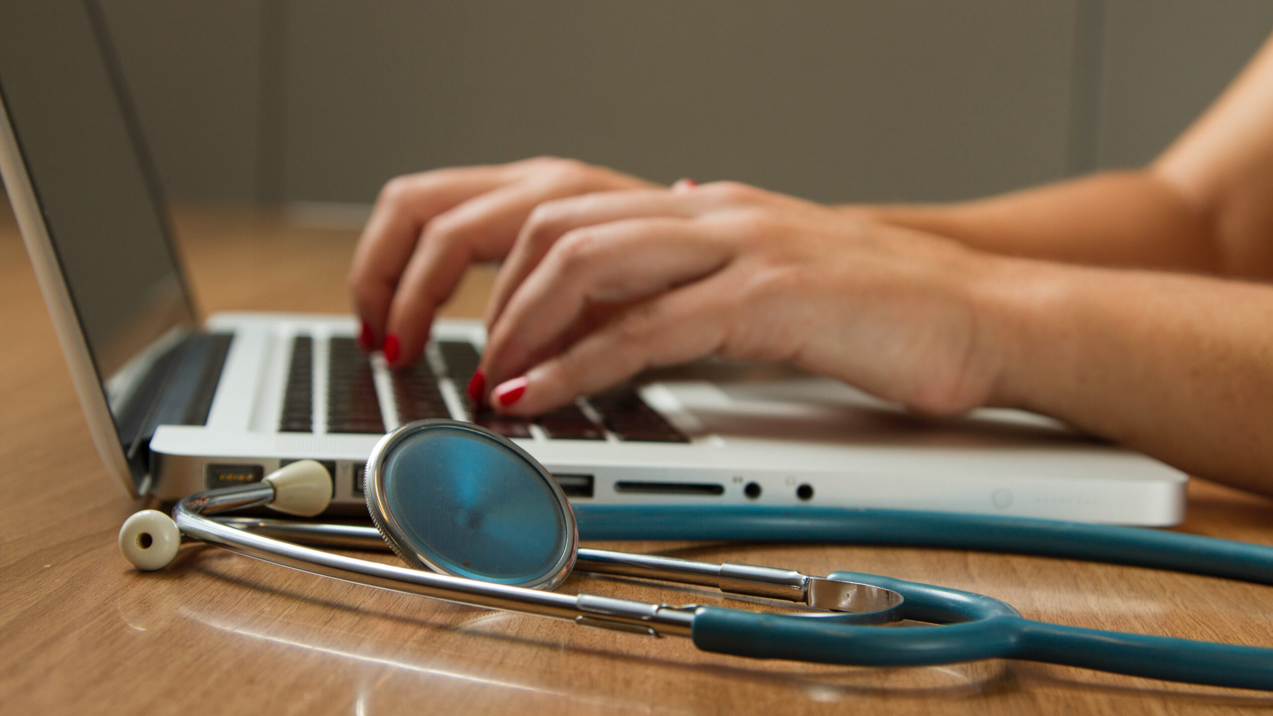 nurse typing on laptop, with stethoscope next to her