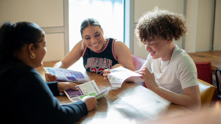 three young people in a Brook workshop, looking at worksheets and smiling