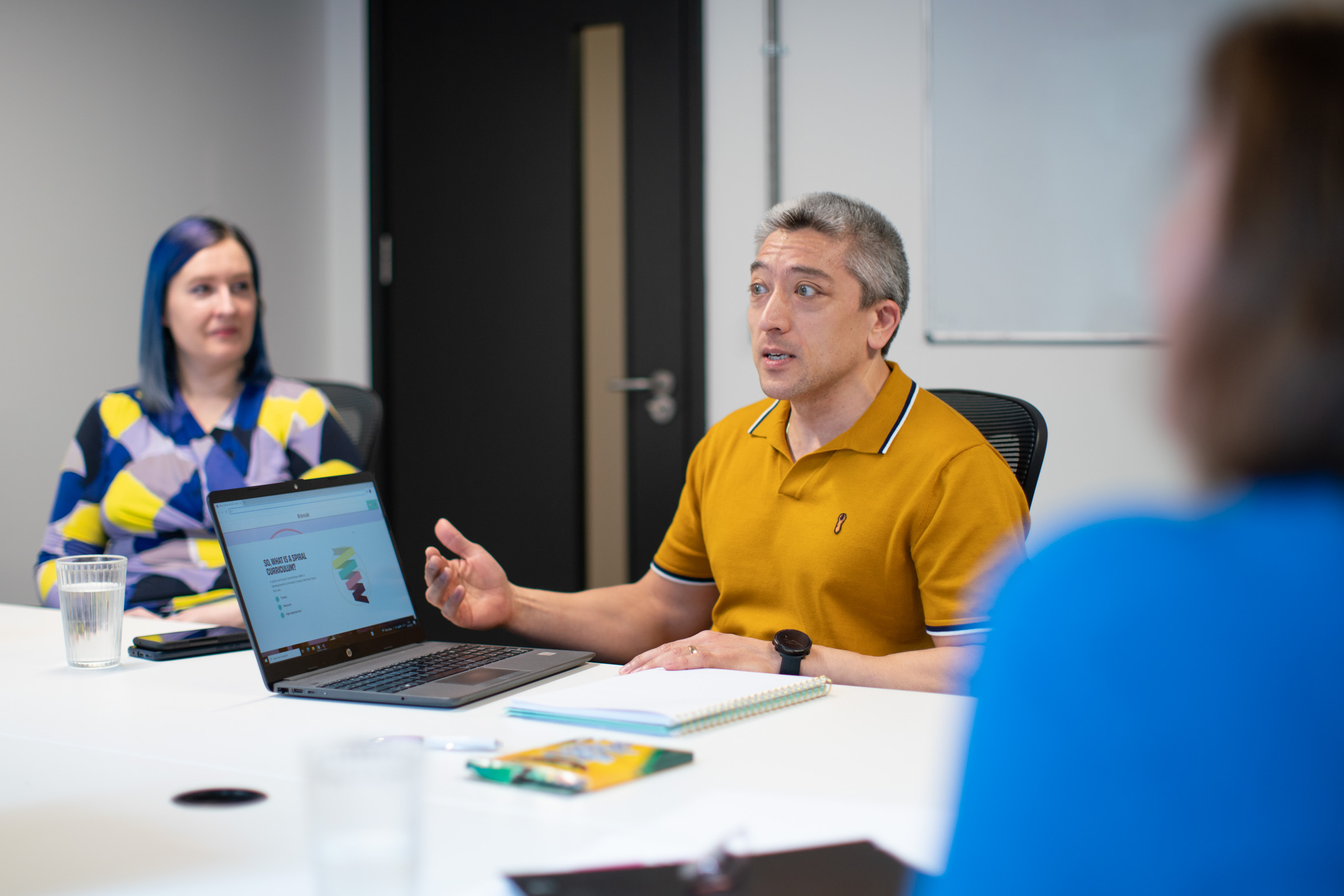 A man talking to other professionals with a laptop in front of him with a presentation slide reading "what is a spiral curriculum?"