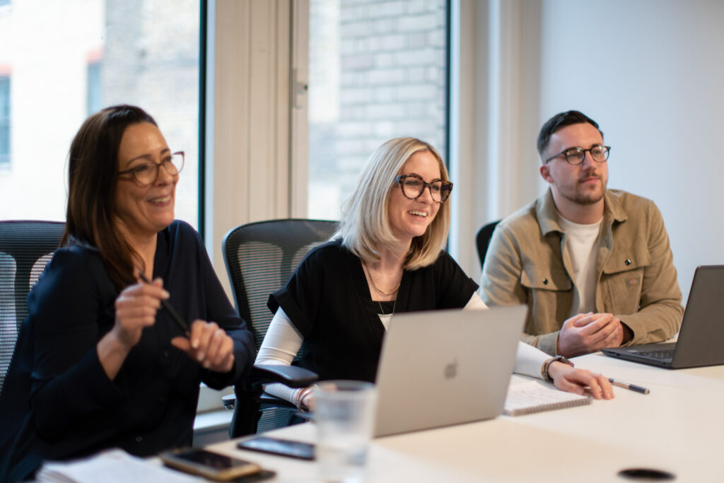 Three professionals with laptops sitting at a desk laughing