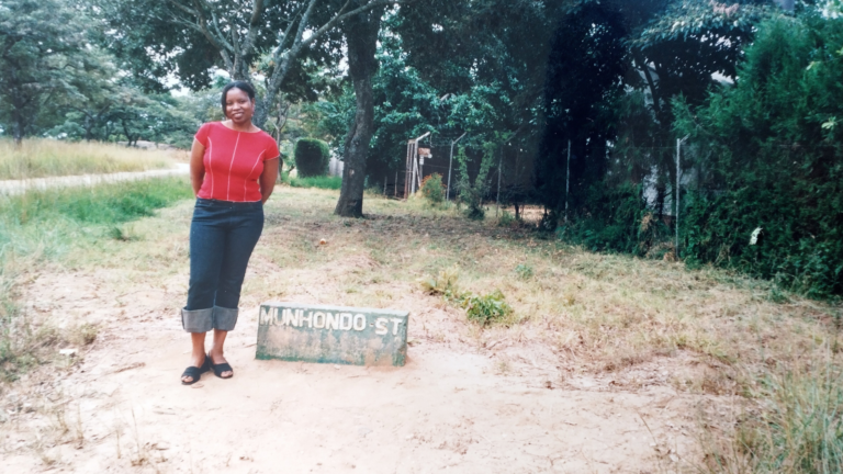 Rutendo standing next to a street sign in Zimbabwe. The ground is dry and there are lots of trees.