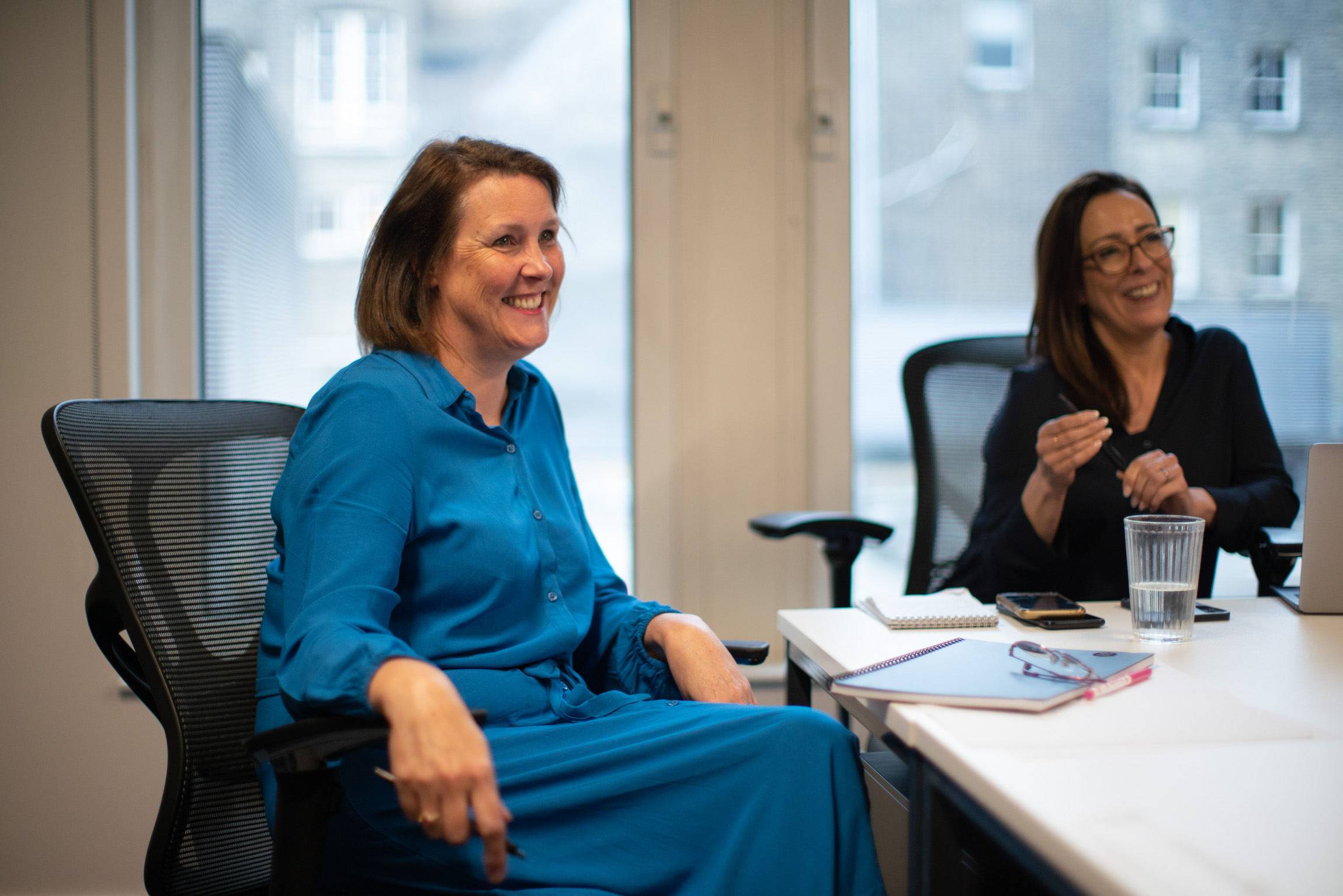 A smily woman is wearing a long blue dress sitting in a board room setting with a notepad and glasses on the desk in front of her.