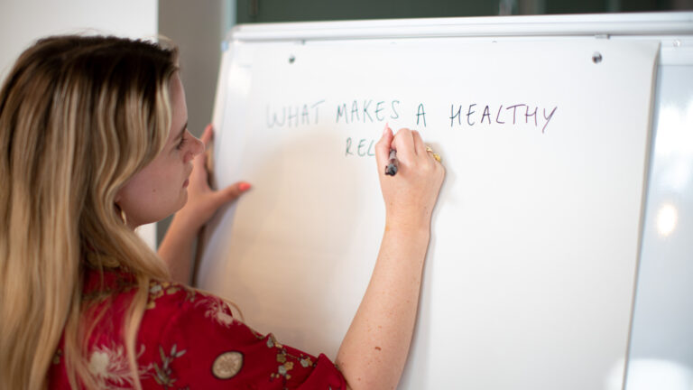 A woman in a red top writes on a whiteboard "what makes a healthy relationship"