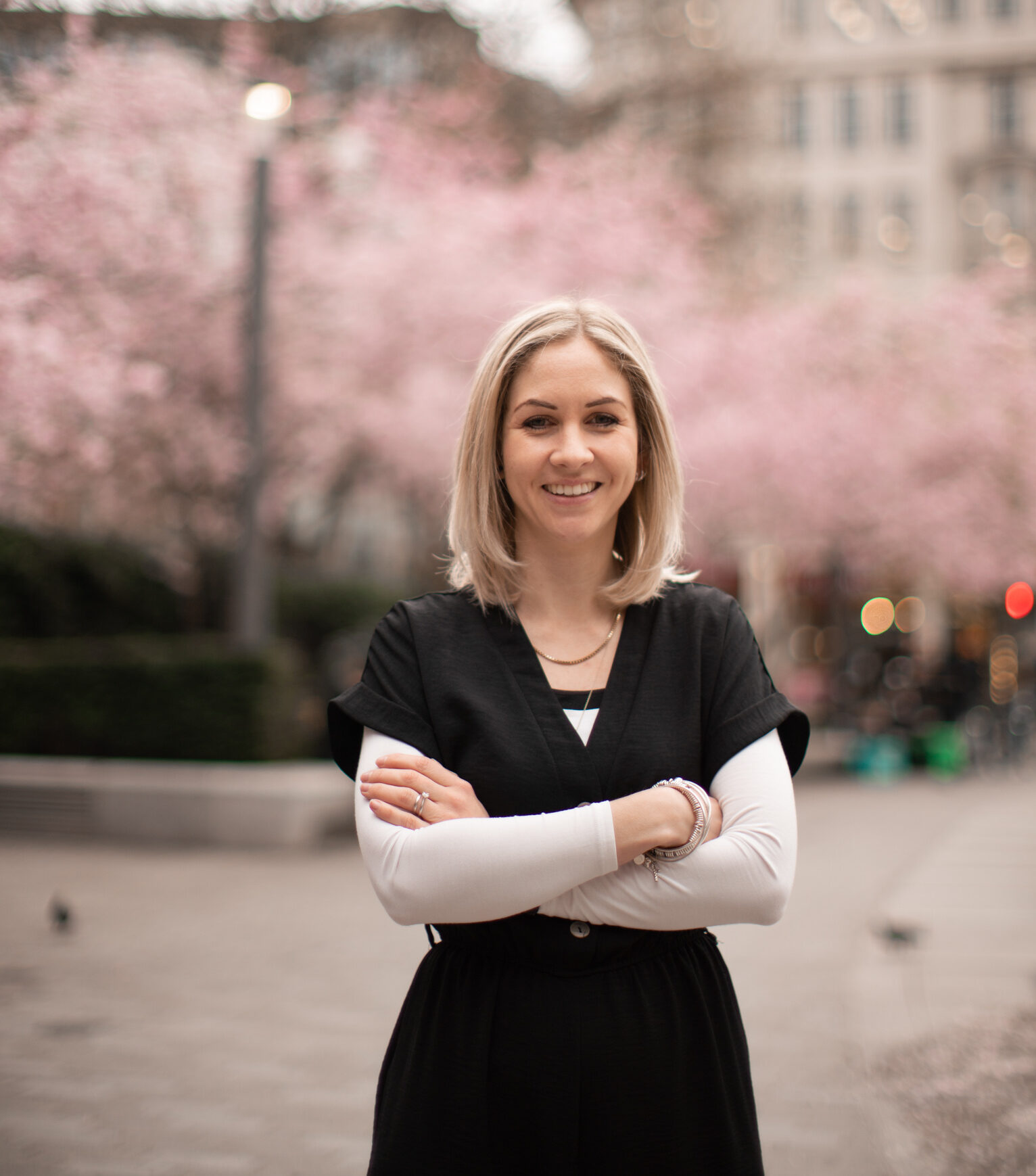 Woman stood with her arms crossed outside with blossom behind her