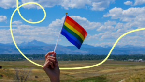 A hand waving an LGBT+ flag with fields in the background