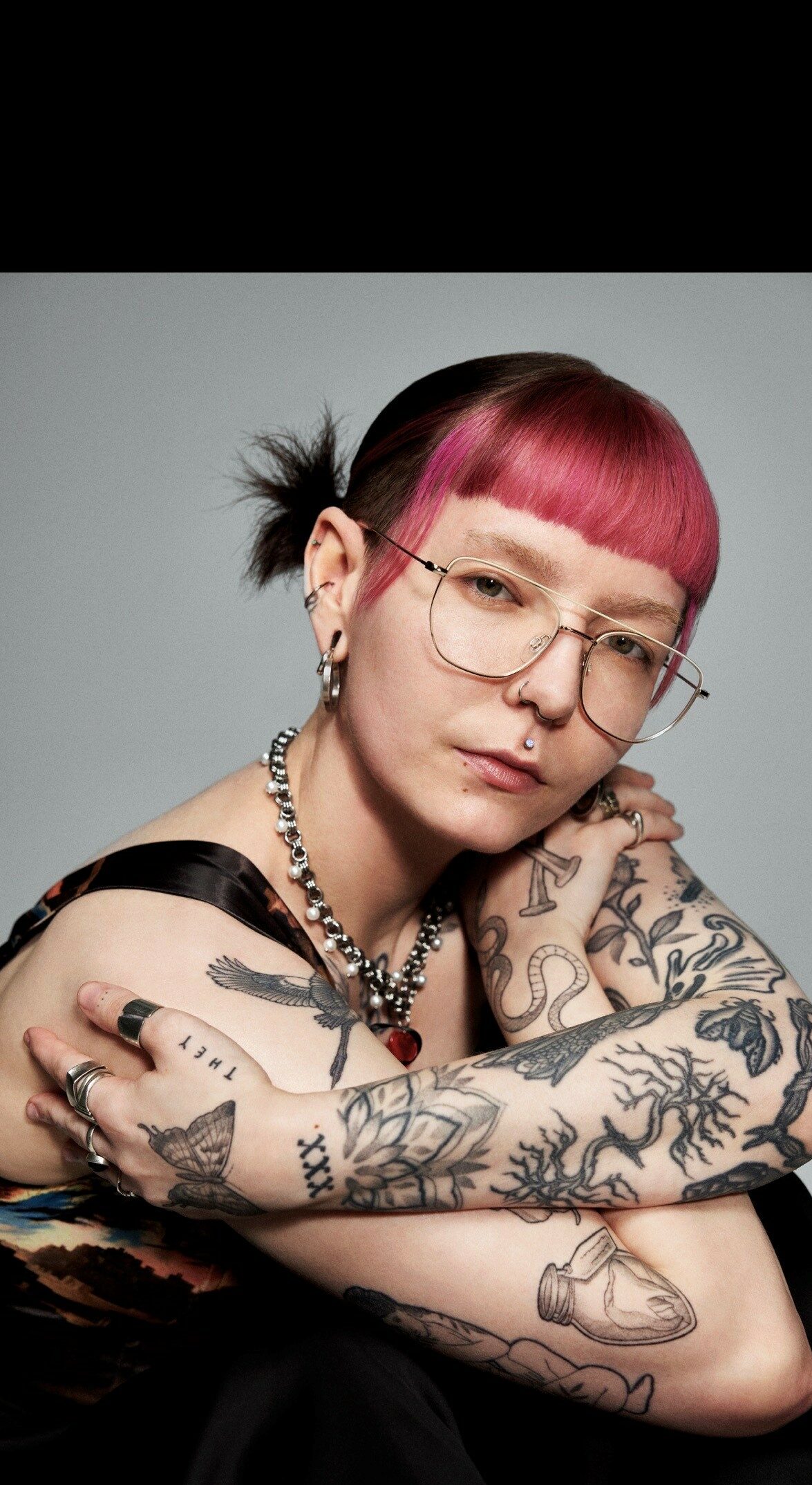Dee wearing glasses and a gold chain necklace with a pink fringe and tattoos
