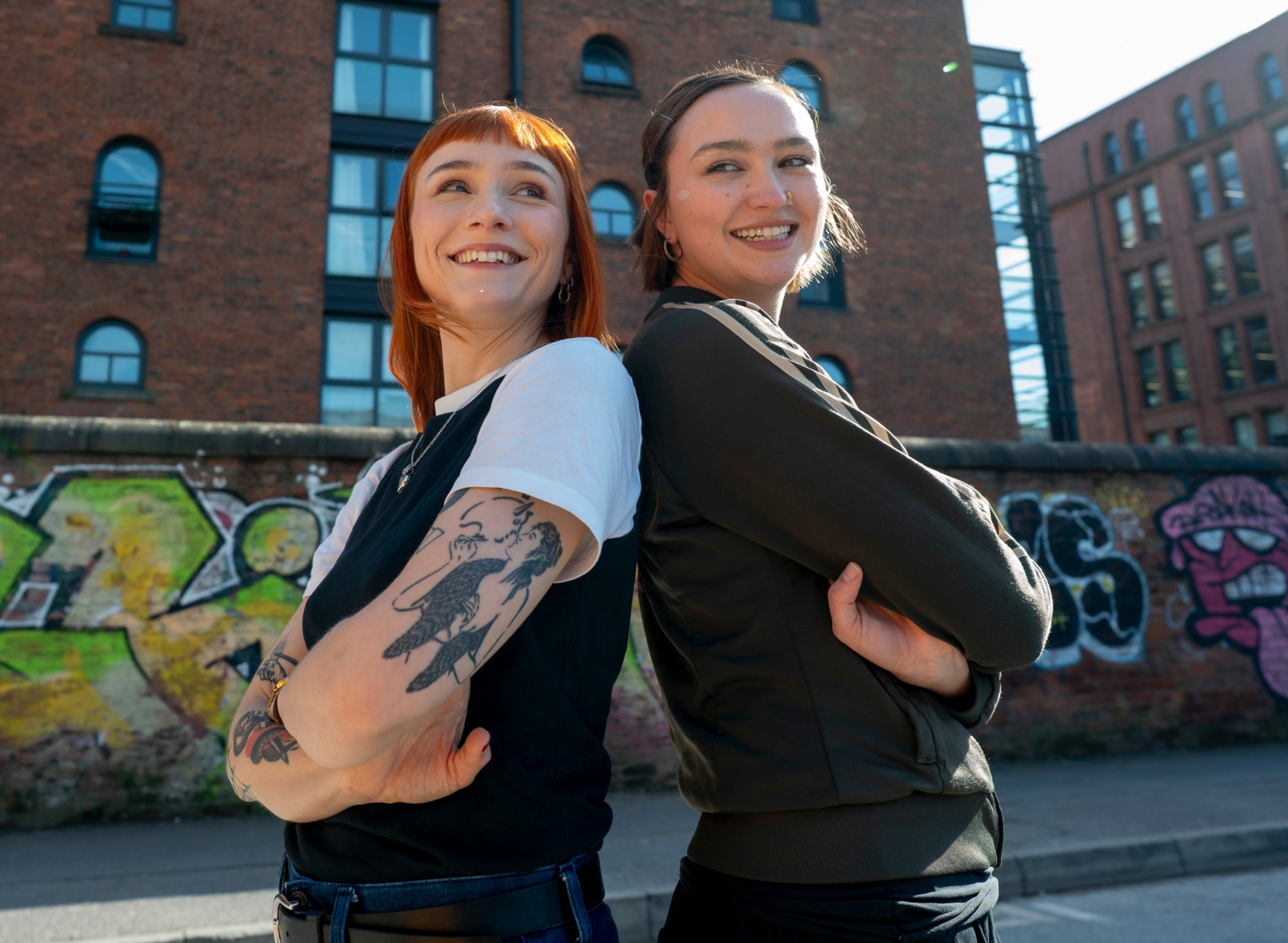 Two young people smiling stood back to back with their arms crossed