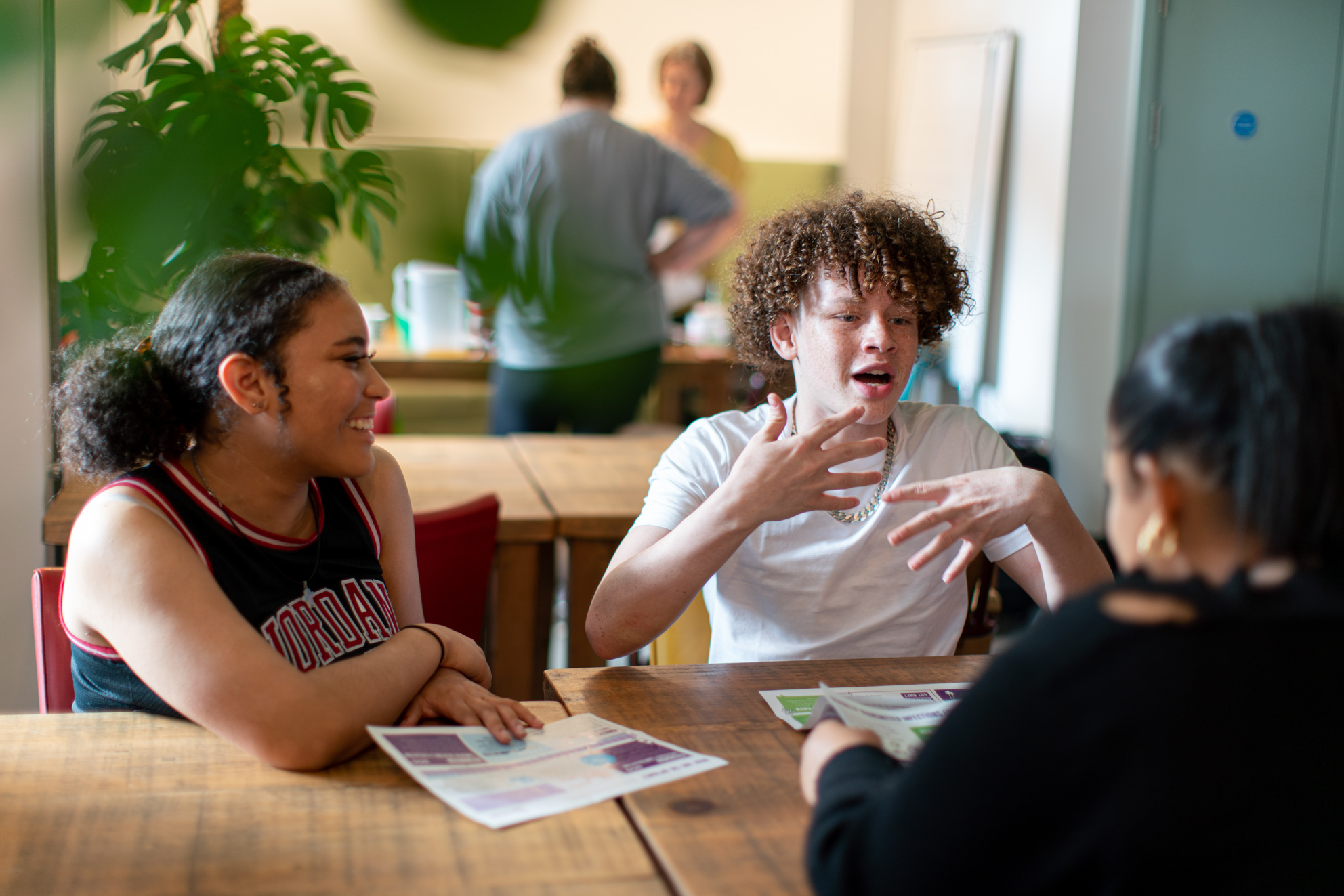 Three students sit around a table, one is talking, one is smiling, and one is facing the speaker but with their back towards the camera.