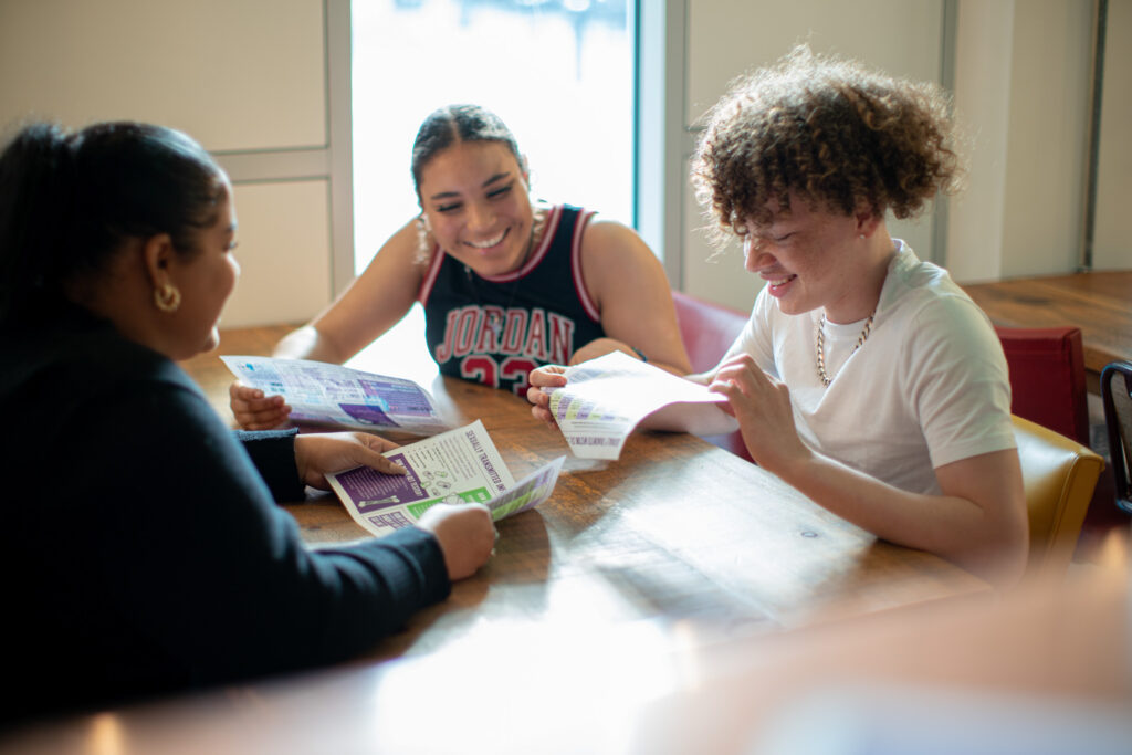 three teenagers sit around a table smiling and looking at papers with text and graphics on them