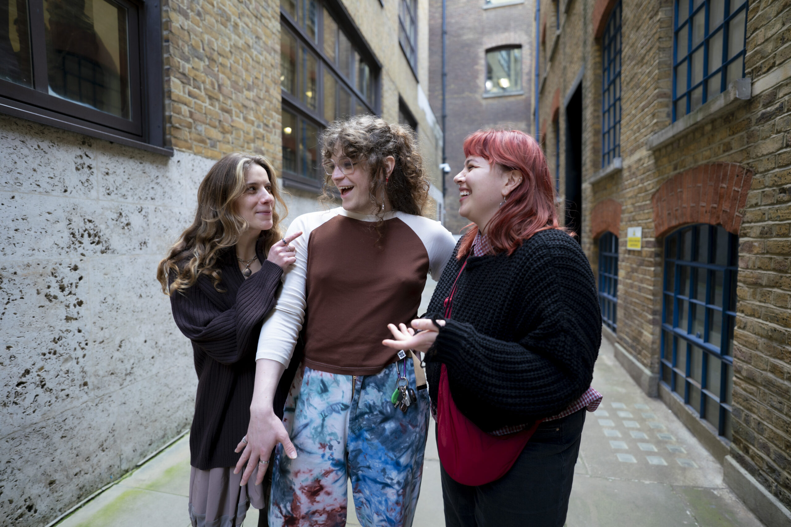 Three people with long hair talk and laugh with each other while walking through an alleyway