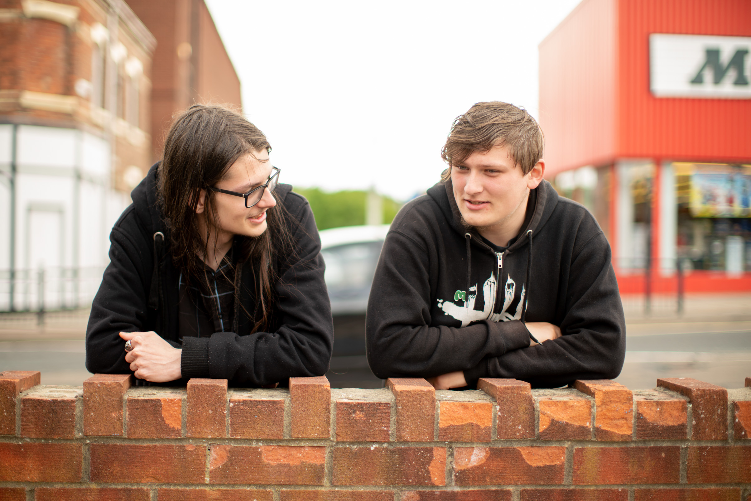 Two young people lean on a brick wall, talking