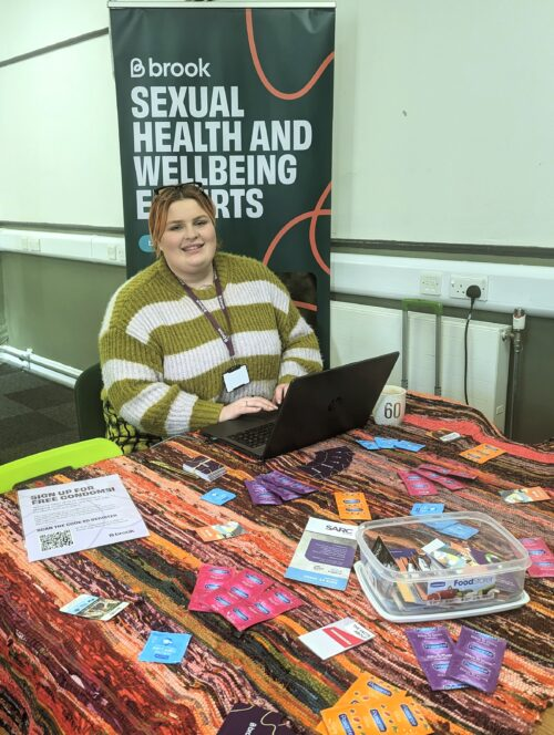 A Brook staff member sits behind a table covered in flyers and packets of condoms
