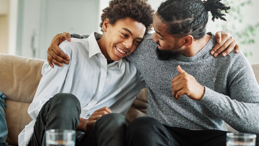 A parent hugs his teenage son. They are sitting on the couch and smiling.