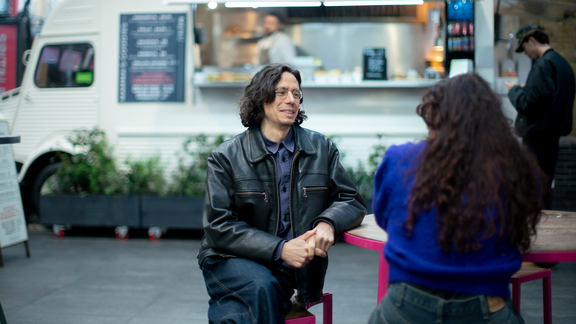 two people sit at a table talking, with a food truck in the background