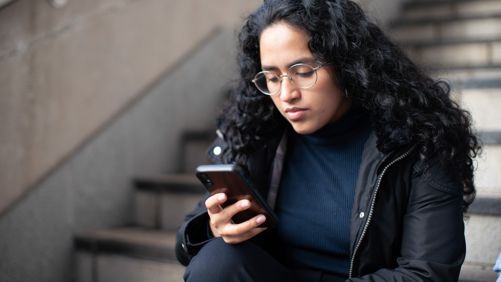 A woman sits on stairs and looks at her phone she is holding.