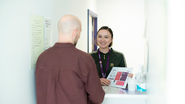 A person at a desk smiles and shows the man in front of the desk a flyer